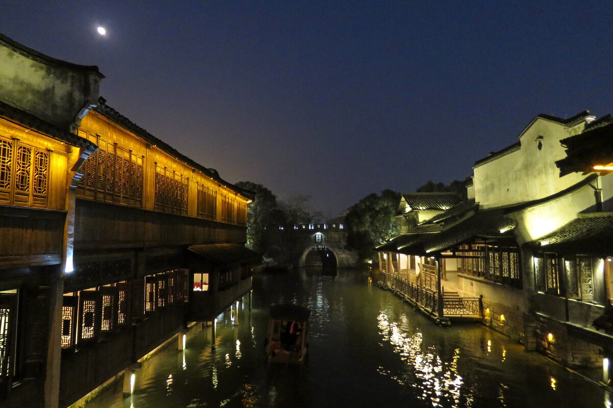Lit-up timber houses along a canal in Wuzhen West Quarter at night, with a moon above and a boat on the water