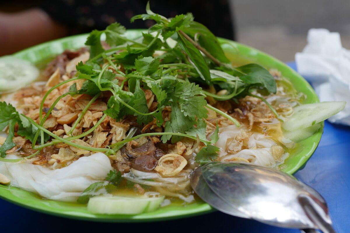 Steamed rice sheets of banh cuon on a green plate, topped with herbs, fried shallots and cucumber