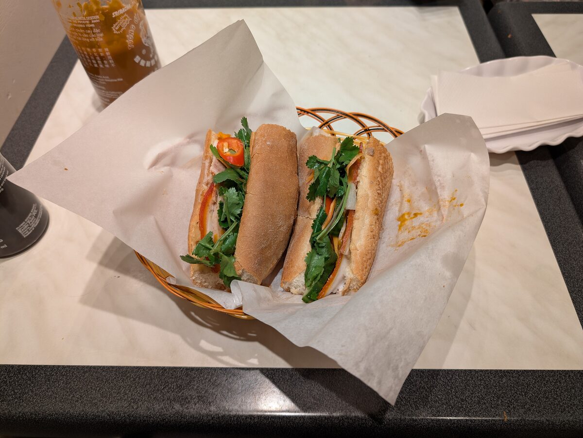 Two baguette sandwiches with herbs and pickles, wrapped in paper on a restaurant table