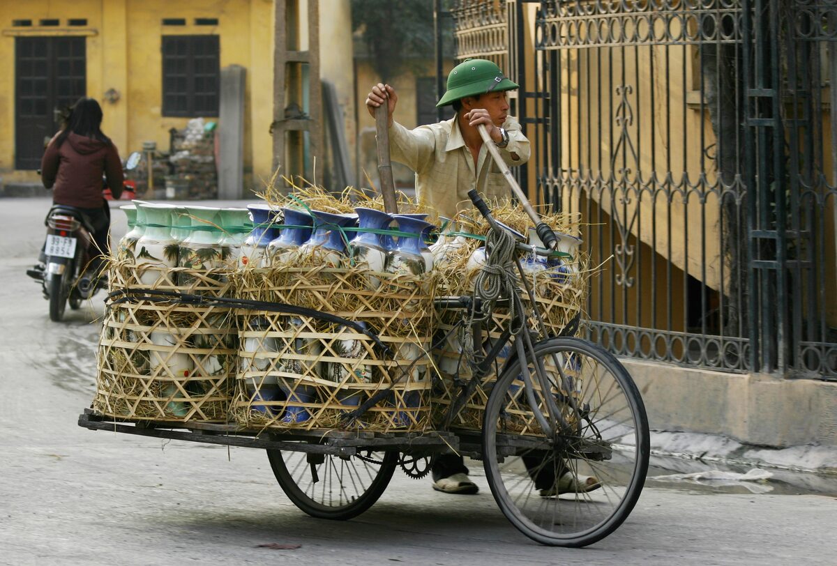 A man pushing a cargo bicycle loaded with blue and white ceramic vases packed in straw baskets along a Bat Trang street