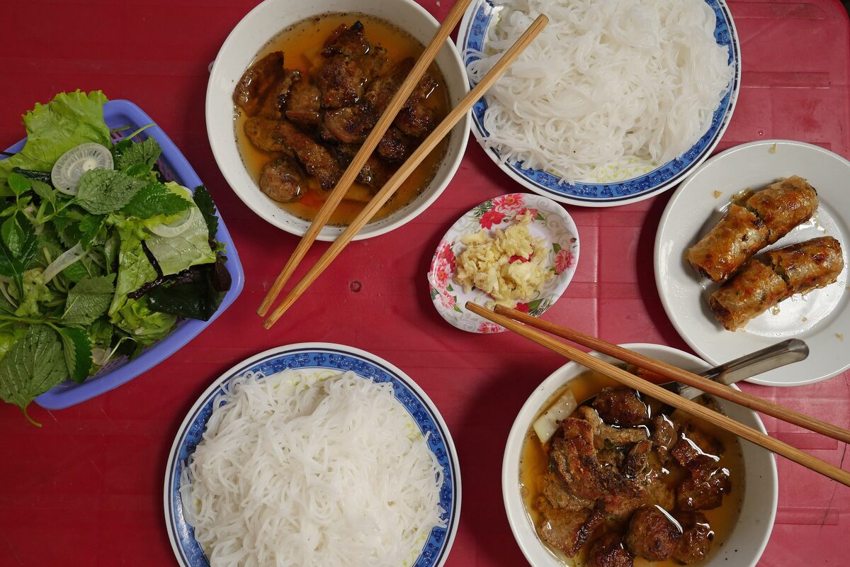 Bun cha on a table: grilled pork in a bowl of broth, rice noodles, a plate of herbs and fried spring rolls