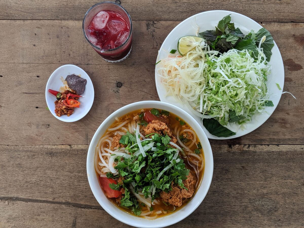 A bowl of bun rieu with tomato, crab and herbs, beside a plate of shredded greens on a wooden table