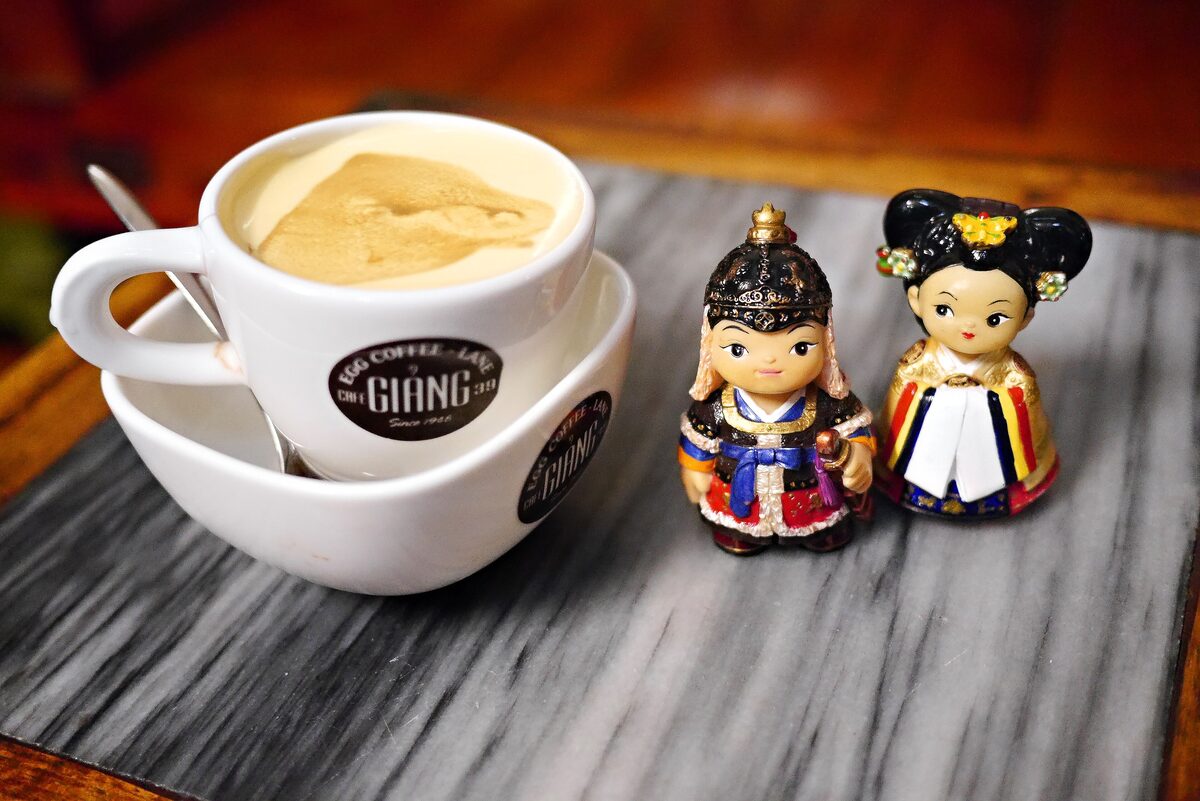 A cup of egg coffee with creamy foam on a saucer from Cafe Giang, next to two small figurines