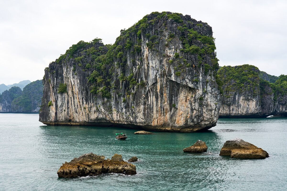 A tall limestone rock rising from the green-blue water of Ha Long Bay, with smaller rocks in the foreground