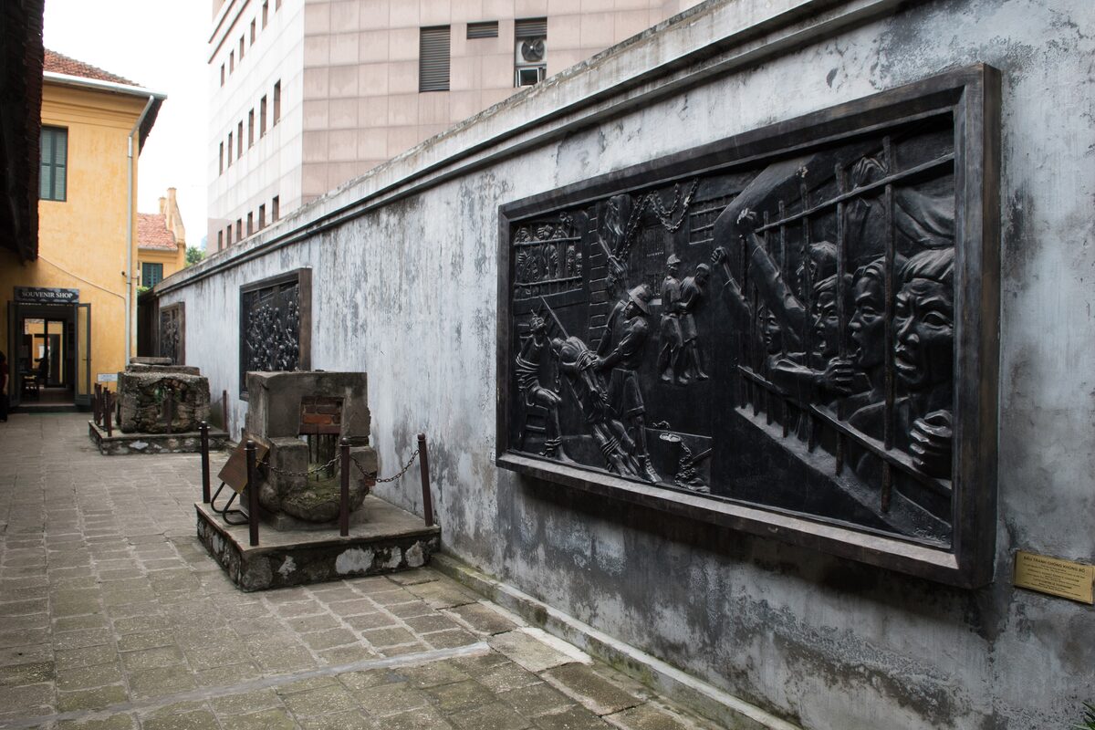 A dark bronze relief of prisoners on a weathered grey wall in a courtyard of the Hoa Lo Prison museum, with stone relics beside it