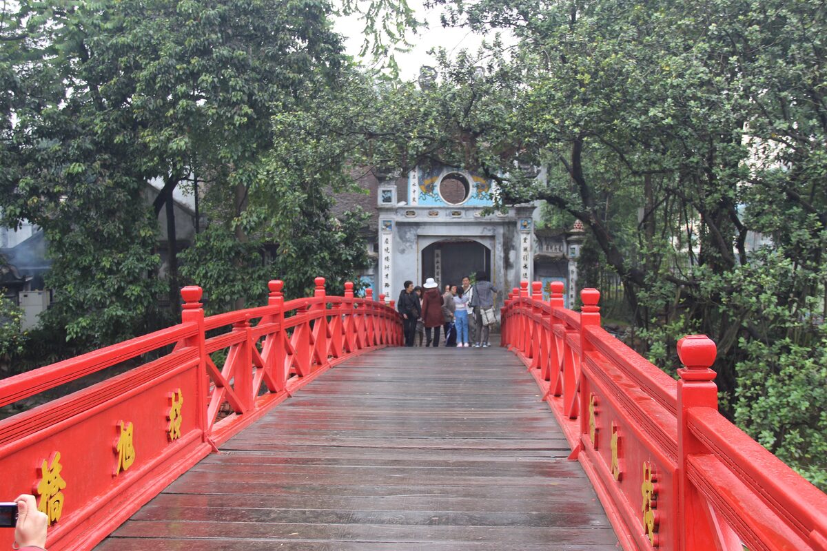 The red Huc Bridge leading through trees to the gate of Ngoc Son Temple, with a few visitors on the bridge