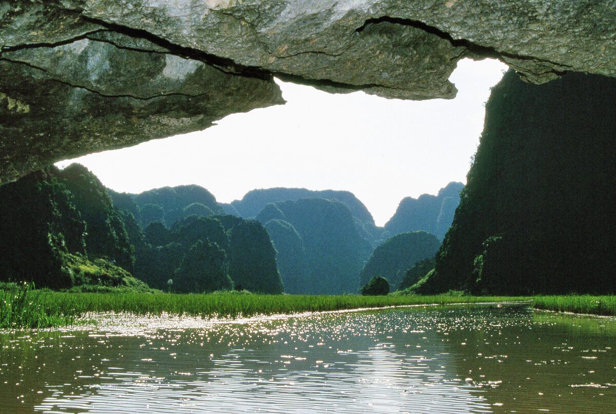 A river in Ninh Binh seen from under a rock overhang, with green karst towers and reeds beyond