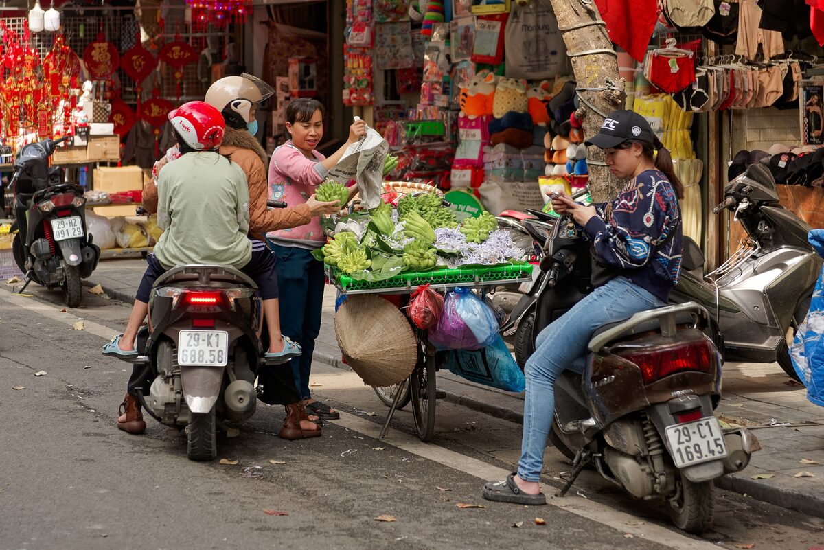 A street vendor's cart of fresh greens among scooters on a busy Old Quarter street, with red shop decorations behind