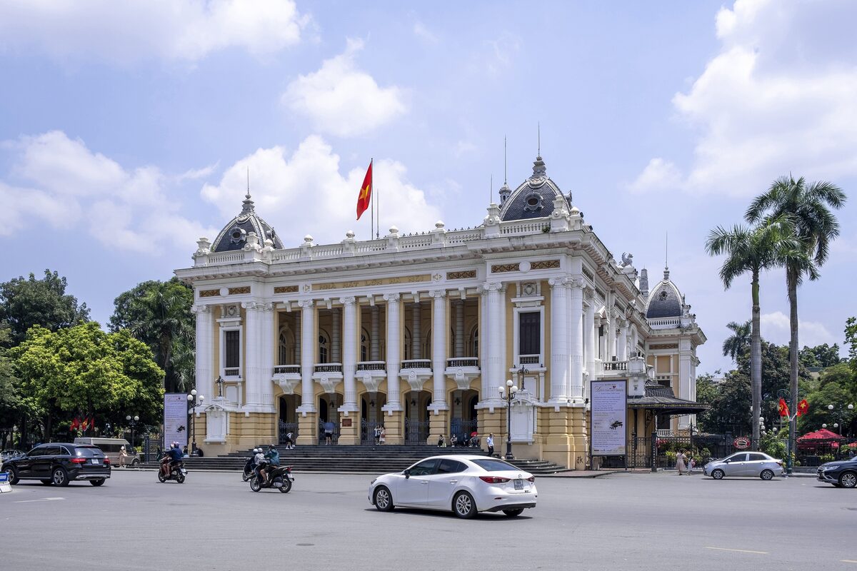 The white colonial facade of the Hanoi Opera House on a sunny day, with palm trees and cars in front