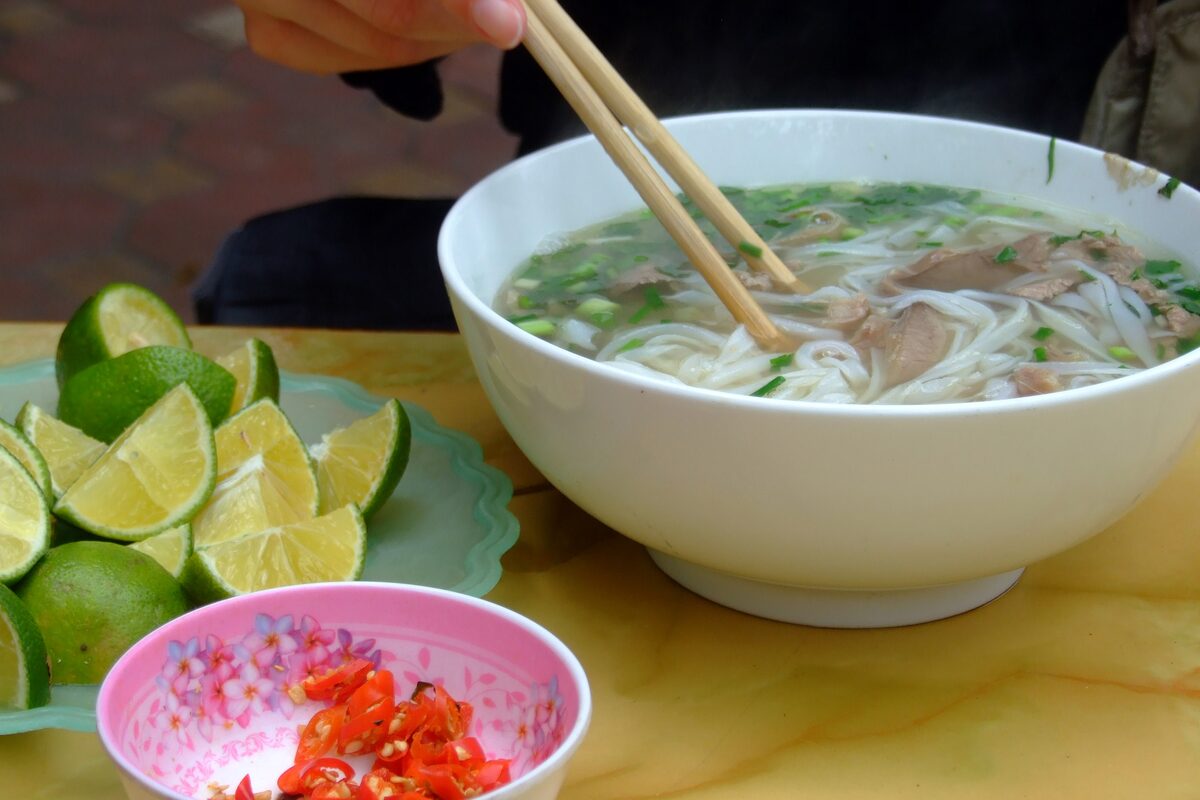 A bowl of beef pho with rice noodles and spring onion, with a plate of lime wedges and a dish of chopped chili