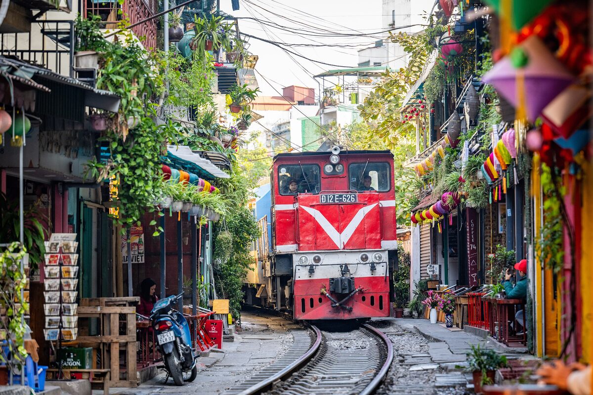 A red train rolling along the track between houses decorated with hanging ornaments on Train Street