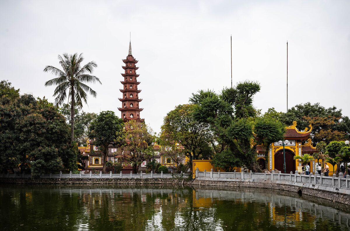 Tran Quoc Pagoda's tall brick tower above the trees beside the lake, with a gate and palm trees