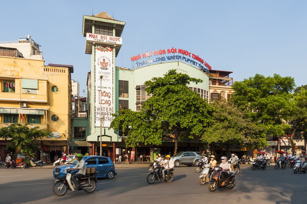 The Thang Long Water Puppet Theatre building with its tall sign tower, seen from a busy street with scooters