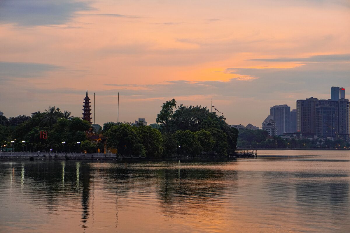 Sunset over West Lake, with the Tran Quoc Pagoda tower among trees on the shore and city towers beyond