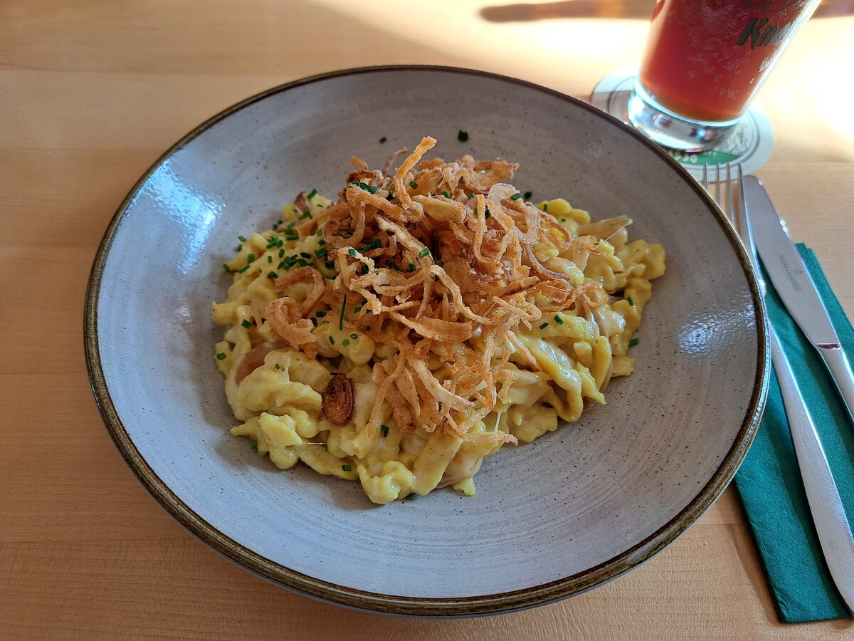 A grey bowl of Käsespätzle topped with crispy fried onions, next to a glass of red drink