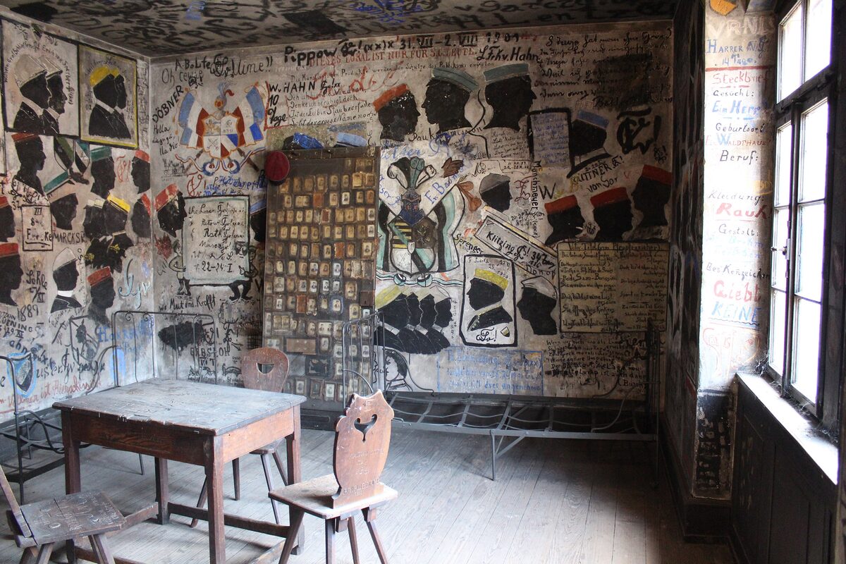 A Studentenkarzer cell with walls and ceiling covered in silhouettes and graffiti, and a table and chairs by the window