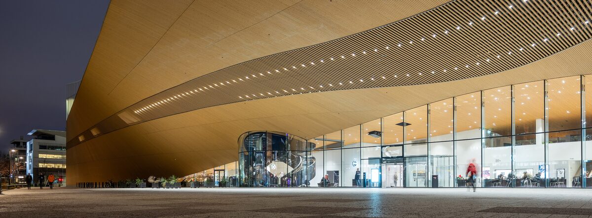 The long, wave-shaped wooden canopy and glass ground floor of Oodi library lit up at dusk