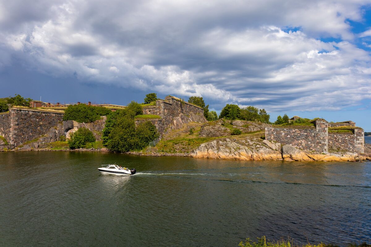 The low stone walls and grassy ramparts of Suomenlinna on its rocky island, with a motorboat passing on the water