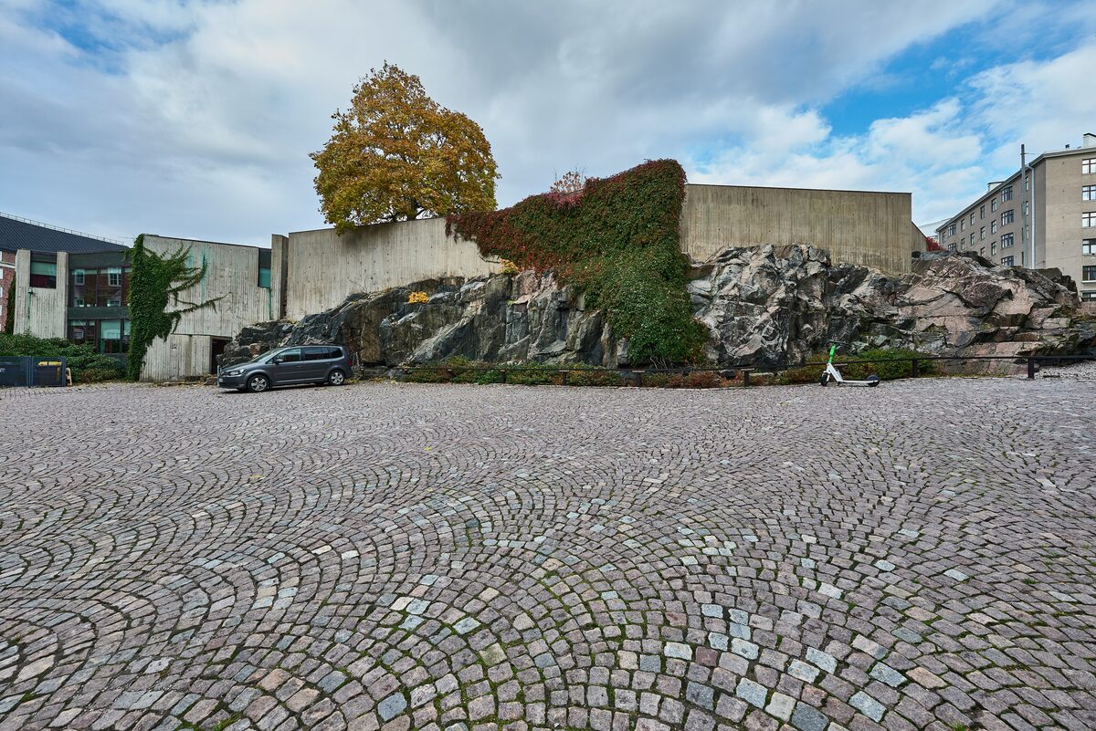 The rough granite outcrop and low concrete walls of the Temppeliaukio Rock Church on a cobbled square
