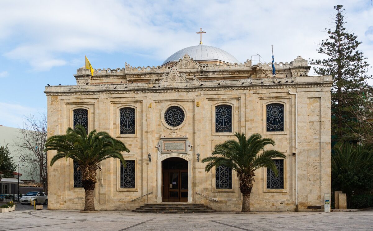 The cream-colored facade of the church of Agios Titos with a white dome, framed by two palm trees