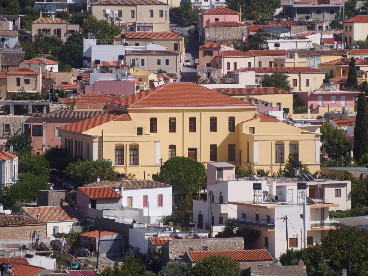 A restored neoclassical building with tall shuttered windows on a square in Archanes, with café chairs in front