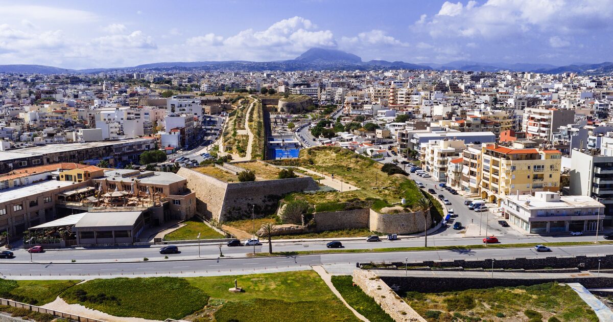 Heraklion from the air, a dense white city running inland behind the green Venetian bastions, with Mount Giouchtas on the horizon