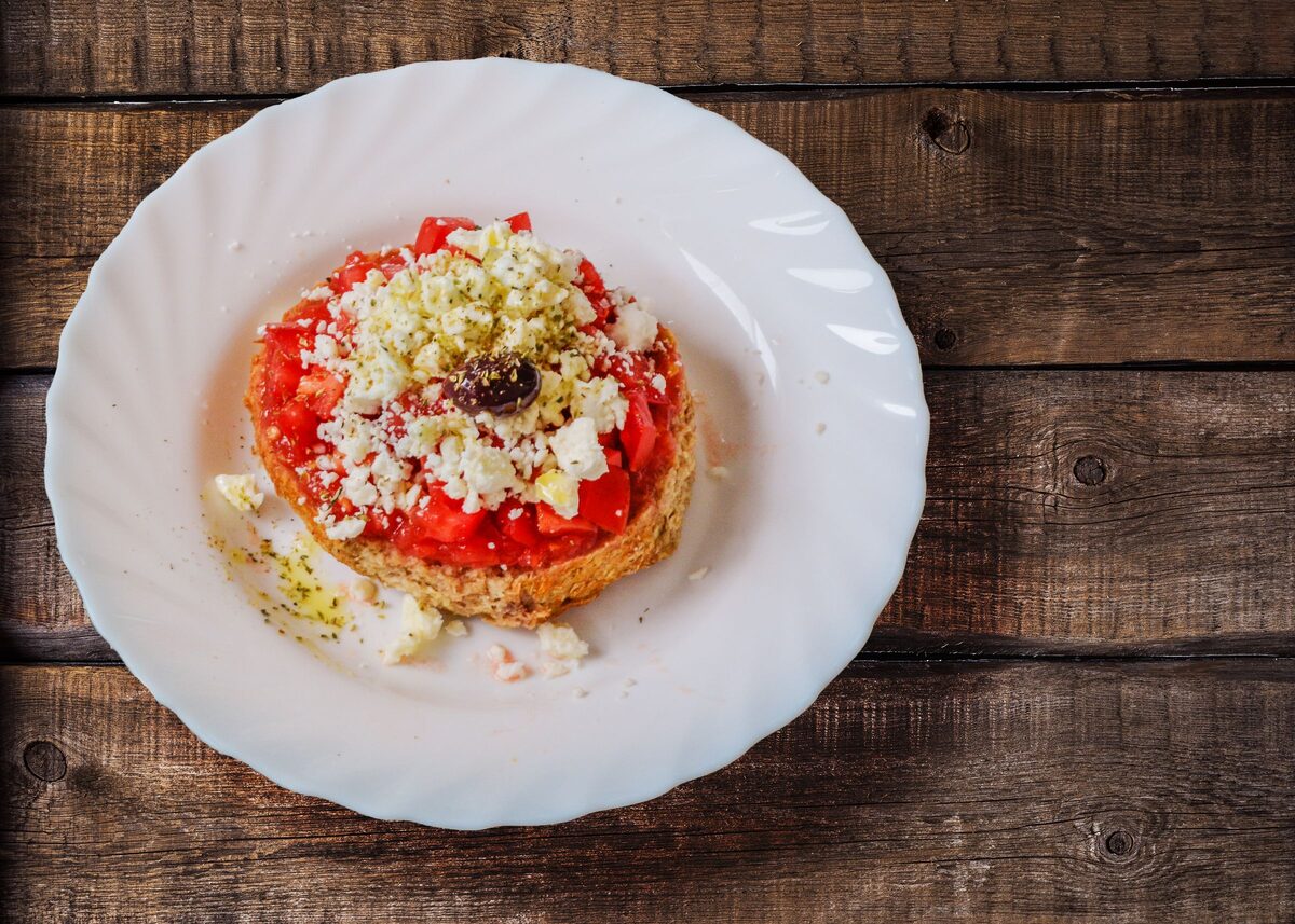 Dakos, a Cretan barley rusk topped with grated tomato, crumbled cheese, an olive and oregano, on a white plate