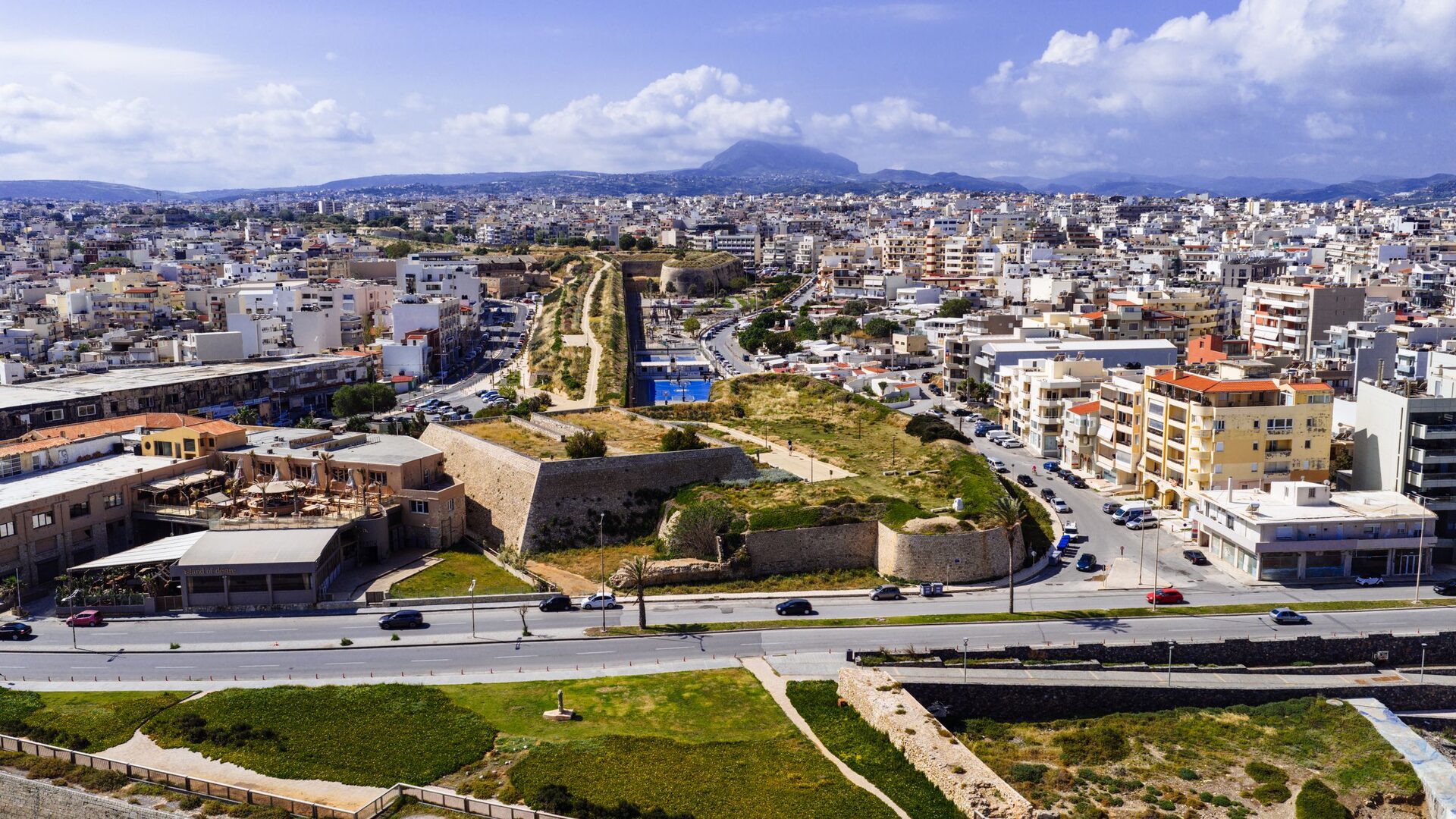 Heraklion from the air, a dense white city running inland behind the green Venetian bastions, with Mount Giouchtas on the horizon
