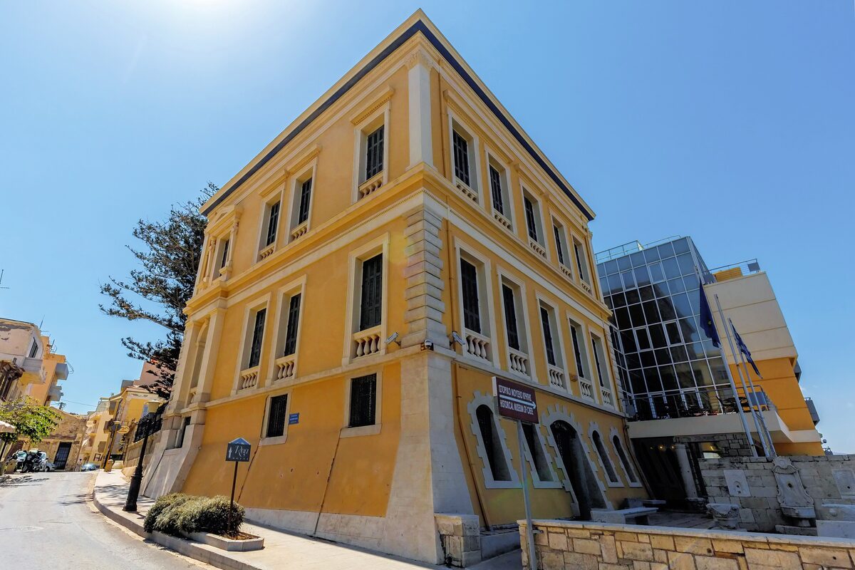 The yellow neoclassical building of the Historical Museum of Crete on a street corner, with a modern glass wing behind it