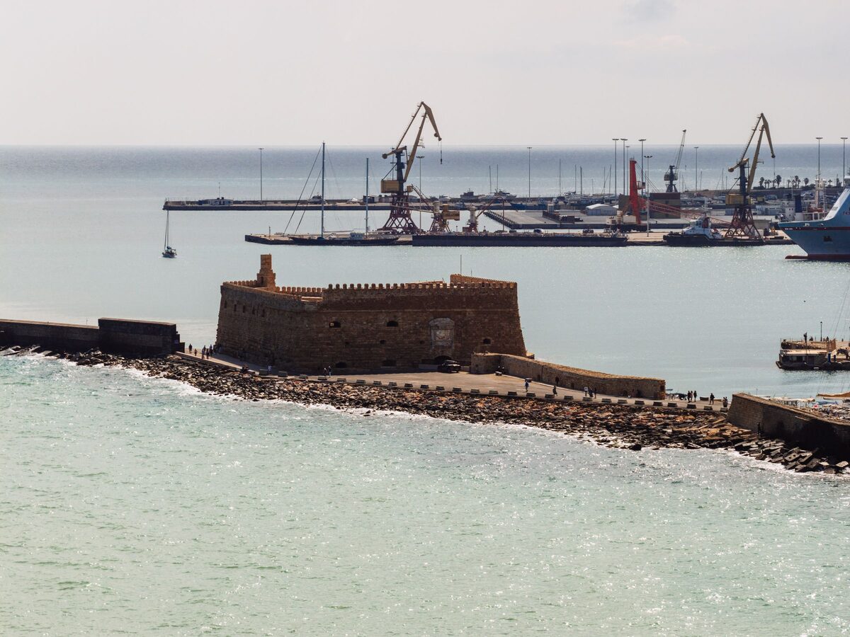 The Venetian fortress of Koules at the end of the harbor breakwater, with calm turquoise water in front and cranes of the port behind