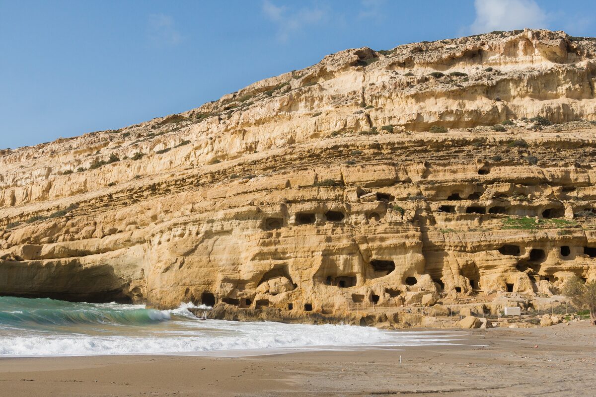 The sandstone cliff of Matala riddled with carved cave tombs, above a sandy beach with waves breaking