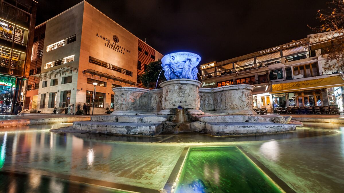 The Morosini fountain with its four stone lions, lit up at night in the middle of Lions Square, with café terraces around it