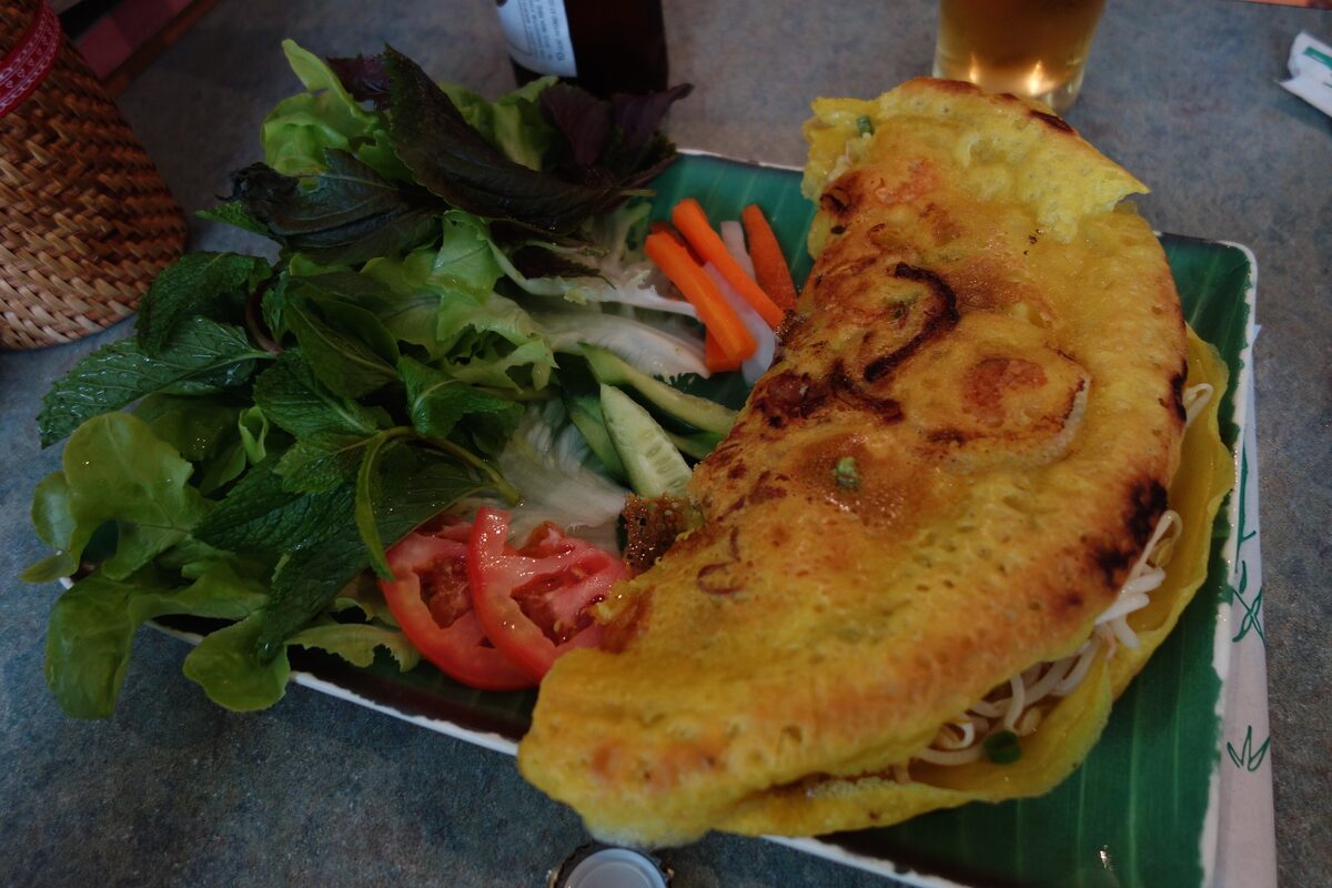 A golden bánh xèo pancake folded over bean sprouts, served on a banana leaf with a pile of herbs, cucumber, carrot and tomato