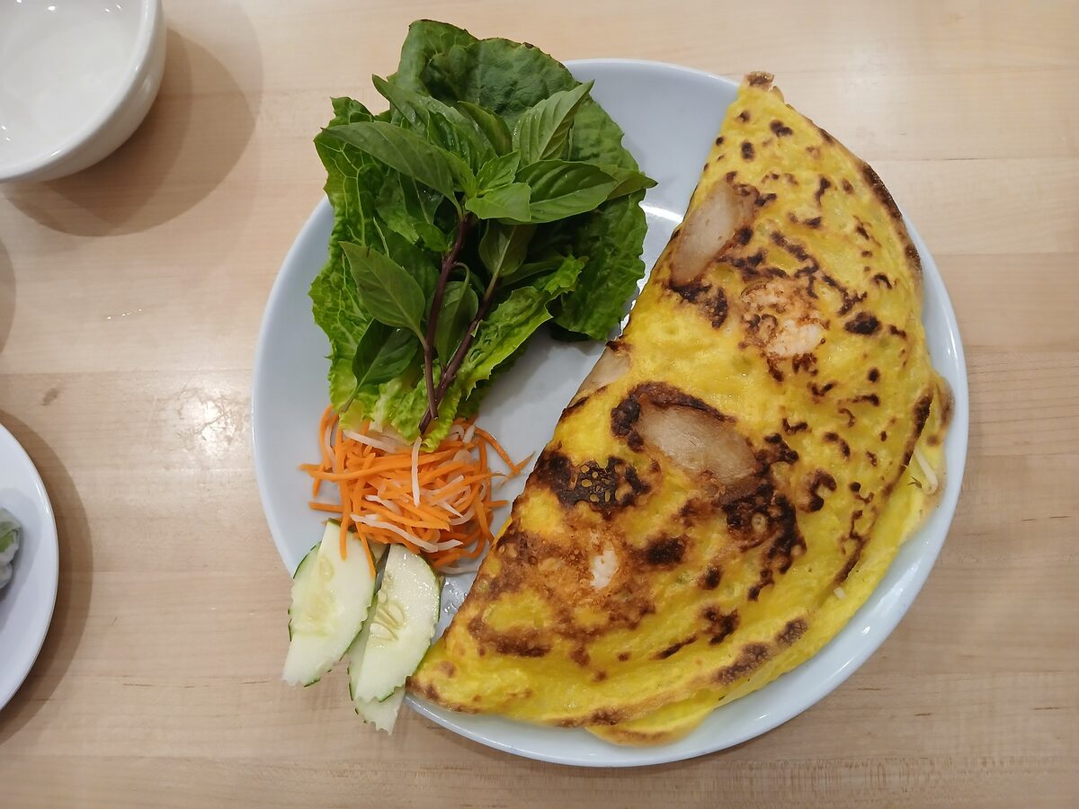 A banh xeo pancake served with pork sausage, shrimp and a plate of fresh garnishes