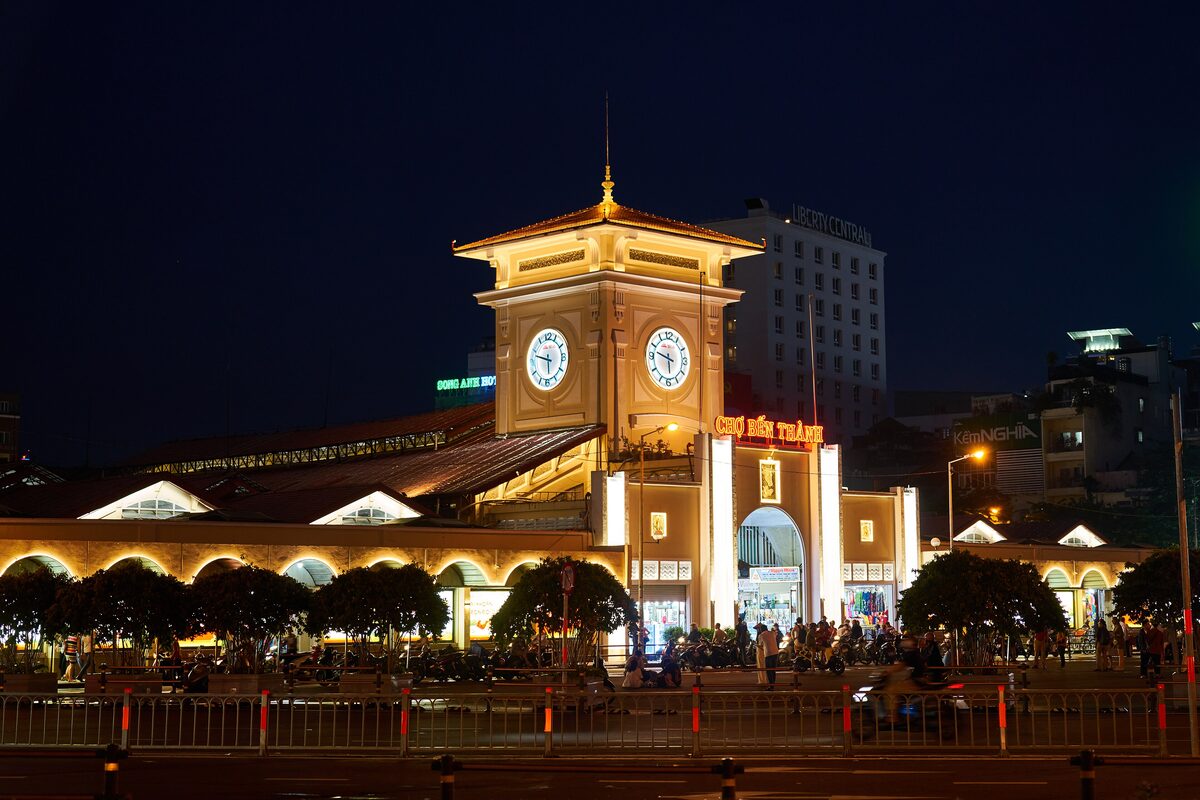 The clock tower and arched gate of Ben Thanh Market lit up at night, with motorbikes and people on the street in front