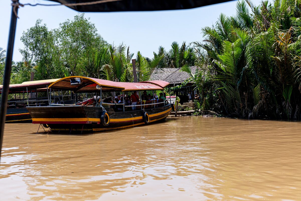A covered wooden tour boat with passengers on a brown river in Bến Tre, with nipa palms and a thatched roof behind