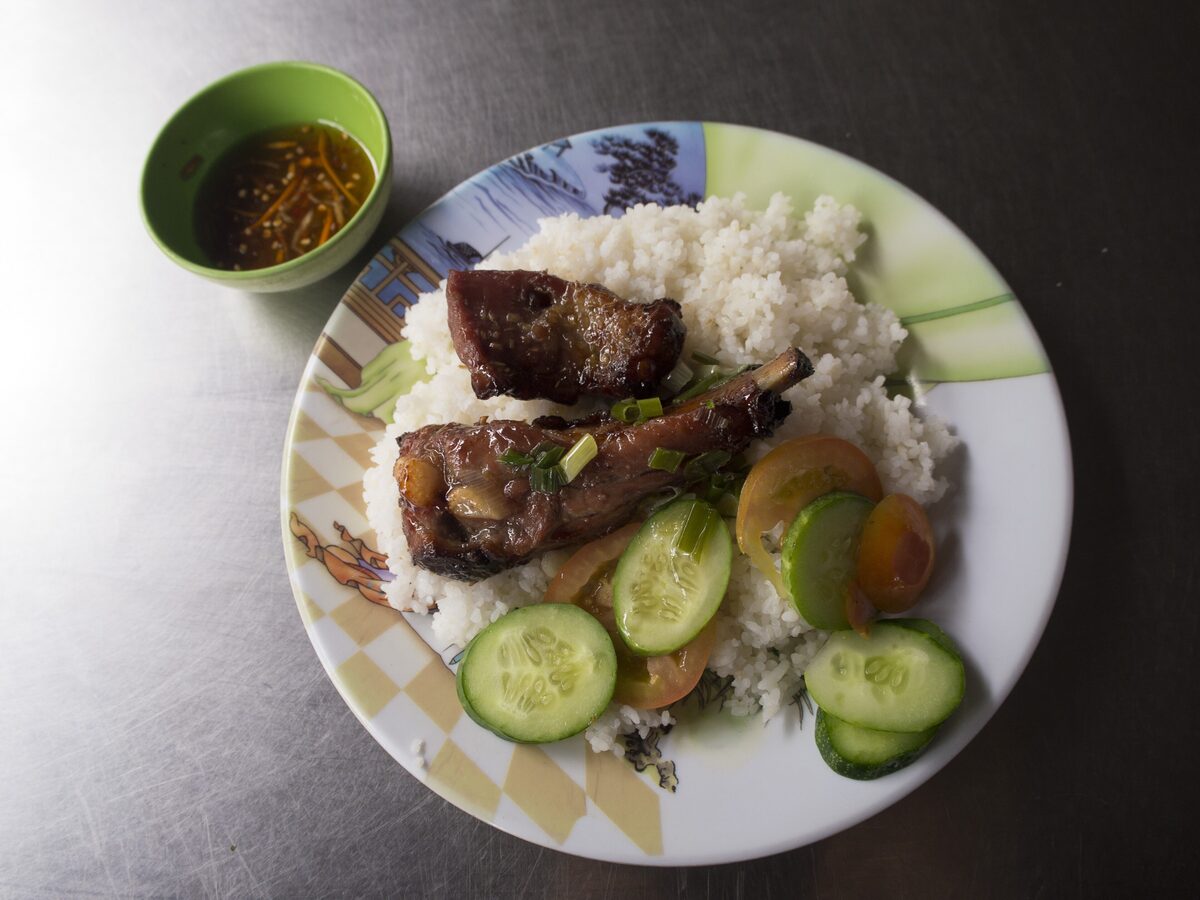 A plate of broken rice with two grilled pork chops, sliced cucumber and tomato, and a small bowl of fish sauce