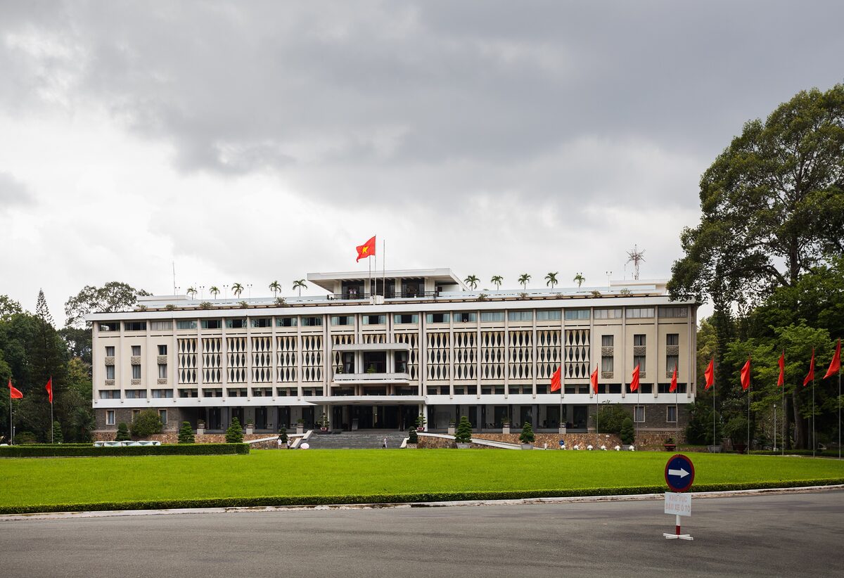 The front of the Independence Palace behind its lawn and flagpoles, with the red flag on the roof