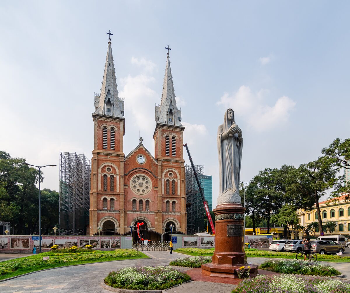 The red-brick Notre-Dame Basilica of Saigon with its twin spires, scaffolding beside the towers and the Virgin Mary statue in front