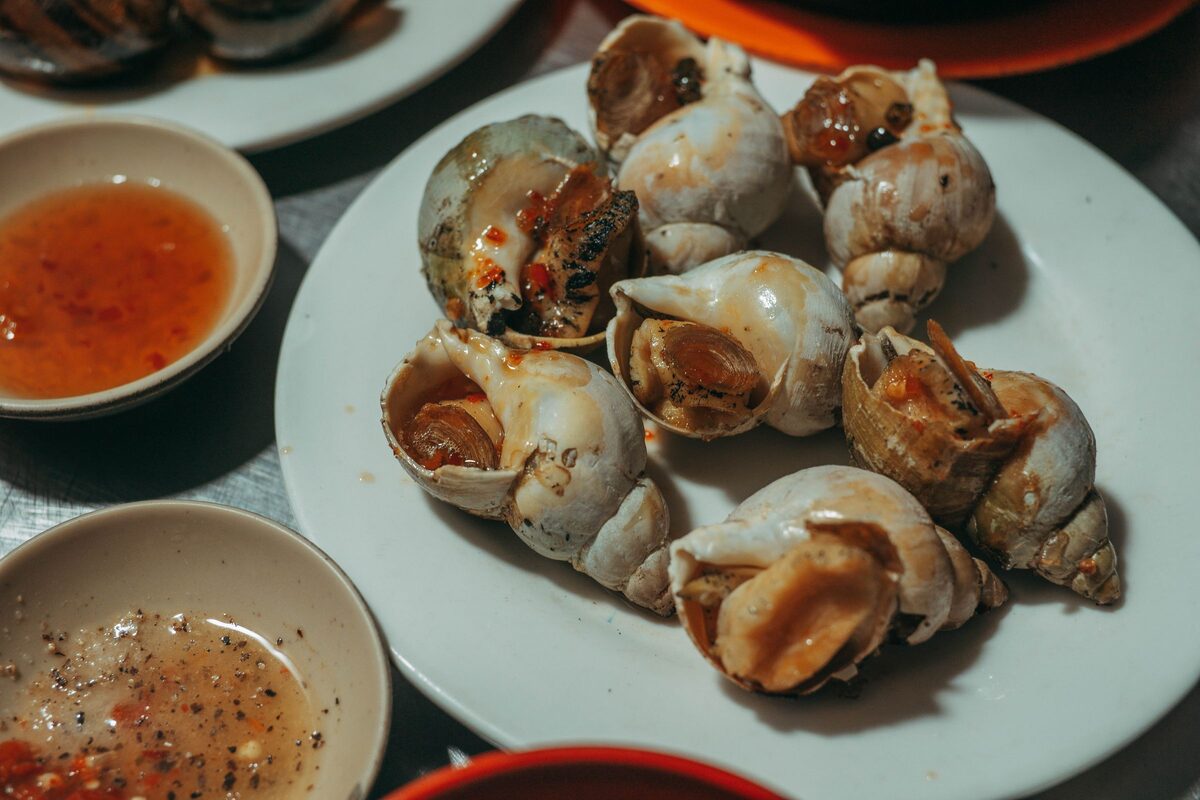 A plate of cooked sea snails in their shells with two small bowls of dipping sauce