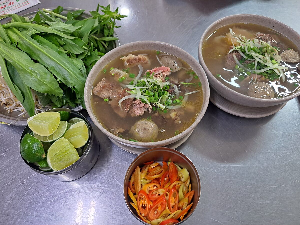 Two bowls of beef pho with herbs, lime and chili laid out on a table