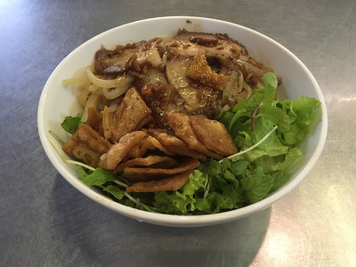 A bowl of cao lau with pork, crisp noodle croutons and lettuce on a steel counter