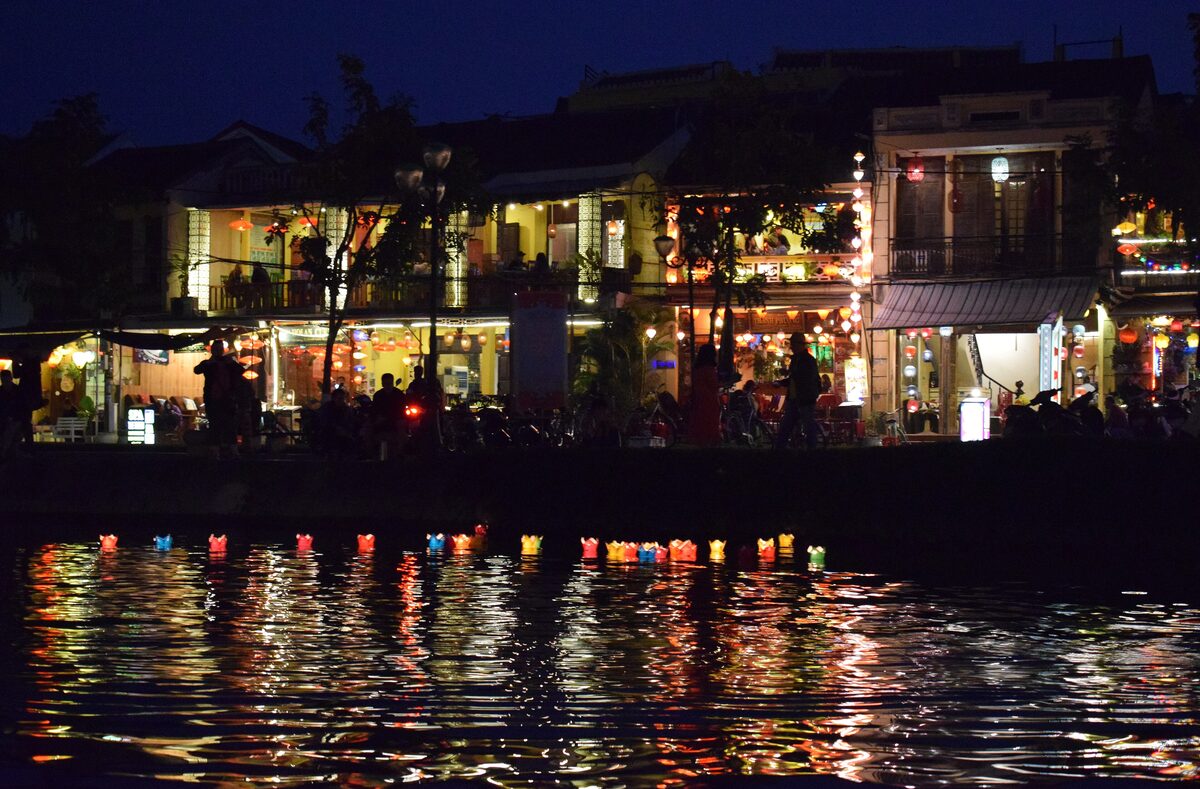 Lanterns float on the Thu Bon at night, reflected in the water beside lit riverside houses