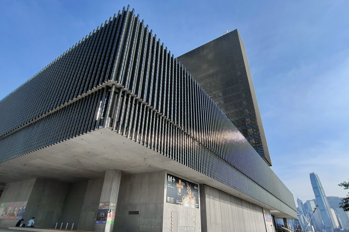 The M+ museum's long ribbed facade and concrete base under a blue sky in West Kowloon