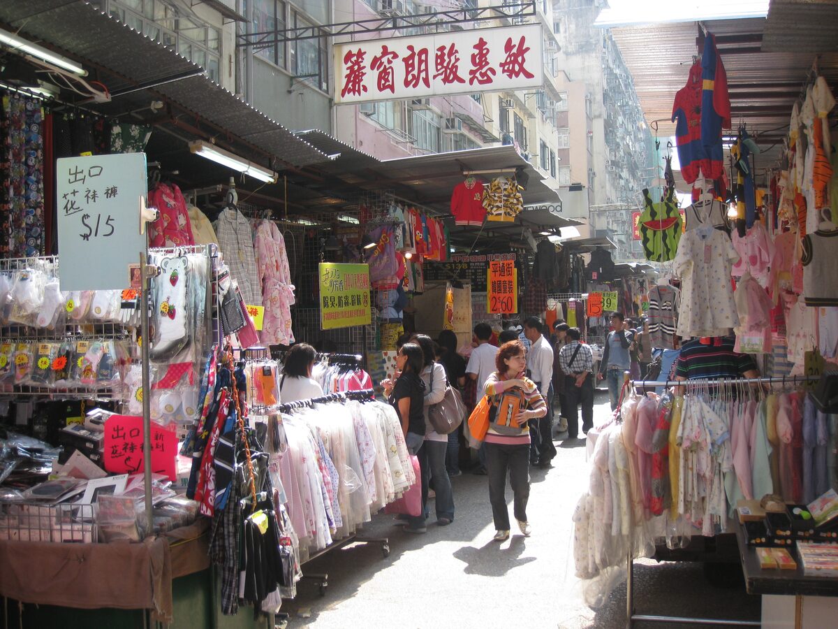 A narrow lane of the Ladies' Market in Mong Kok lined with clothing stalls and awnings, with shoppers walking between the racks