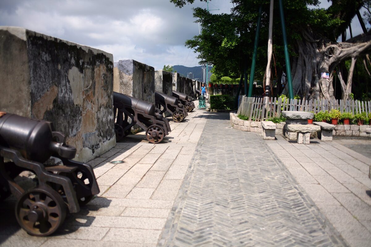 A row of old iron cannons along the stone ramparts of Mount Fortress in Macau, with a paved walkway beside them