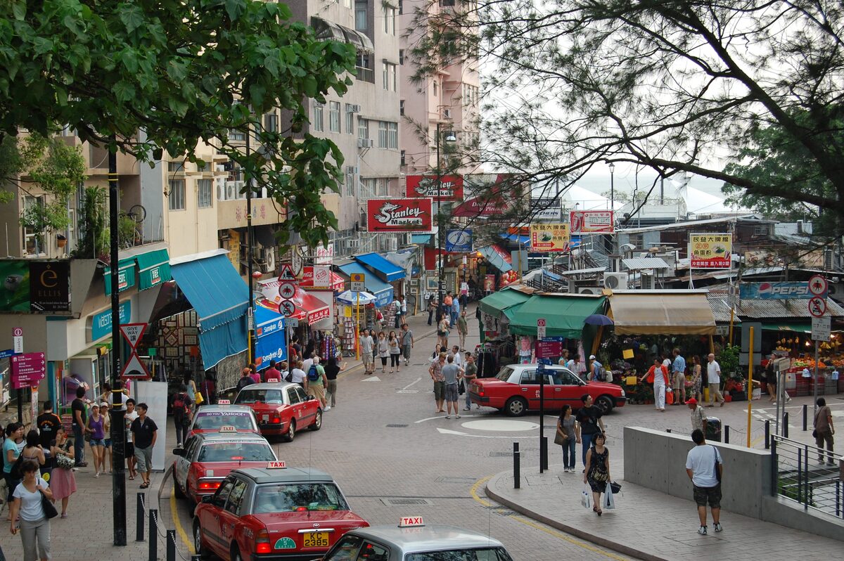 Shoppers and red taxis on the street by Stanley Market, with market awnings and trees along the road