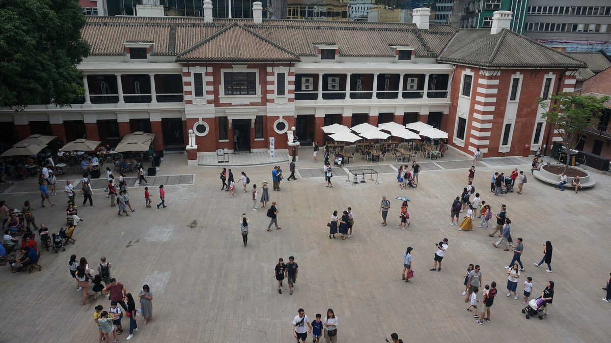 A red-brick and white colonial building of Tai Kwun seen from above, with visitors crossing the open courtyard and cafe umbrellas in front