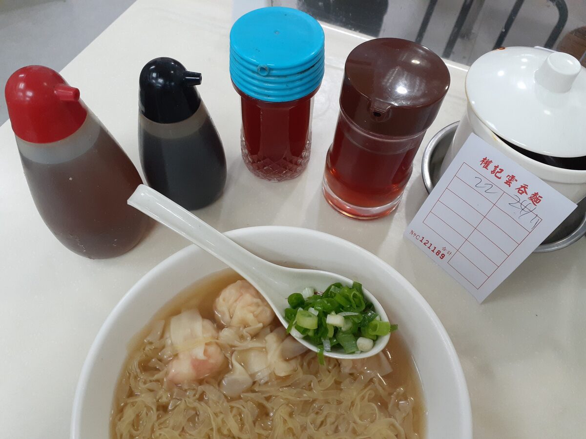 A bowl of wonton noodle soup with shrimp wontons and chopped spring onion, next to sauce bottles on the table