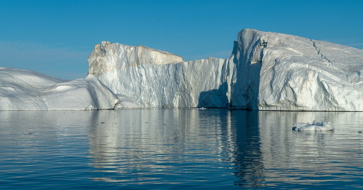 Two white icebergs rising from calm blue water under a clear blue sky, with their reflections in the Ilulissat Icefjord